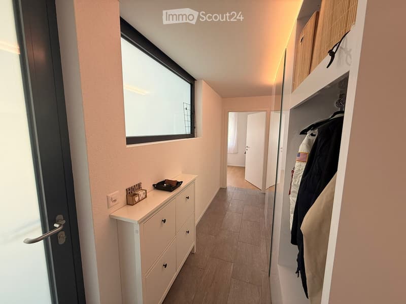 A narrow hallway with light-colored walls, tiled flooring, and a built-in closet with hanging clothes. A small console table with a decorative sign and tray is visible near the entrance.