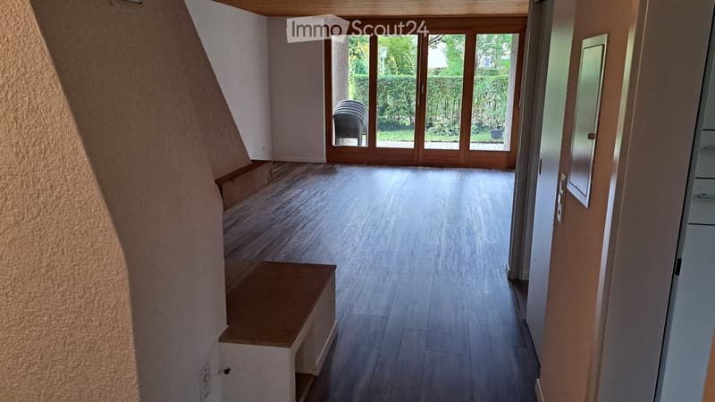 Empty hallway with wooden flooring, large glass doors leading to an outdoor area, and a small bench on the left side.