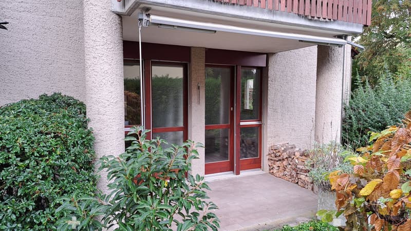 Entrance to a multi-story building with a covered walkway, glass doors, and surrounding greenery.