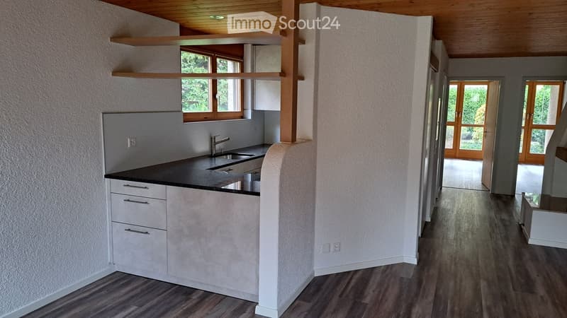Modern kitchen with white cabinets, black countertop, and a sink. The space features wooden flooring and large windows with wooden frames, leading to an open area with wooden doors.