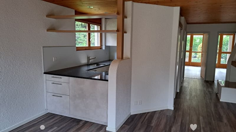 Modern kitchen with white cabinets, black countertop, and a sink. The space features wooden flooring and large windows with wooden frames, leading to an open area with wooden doors.