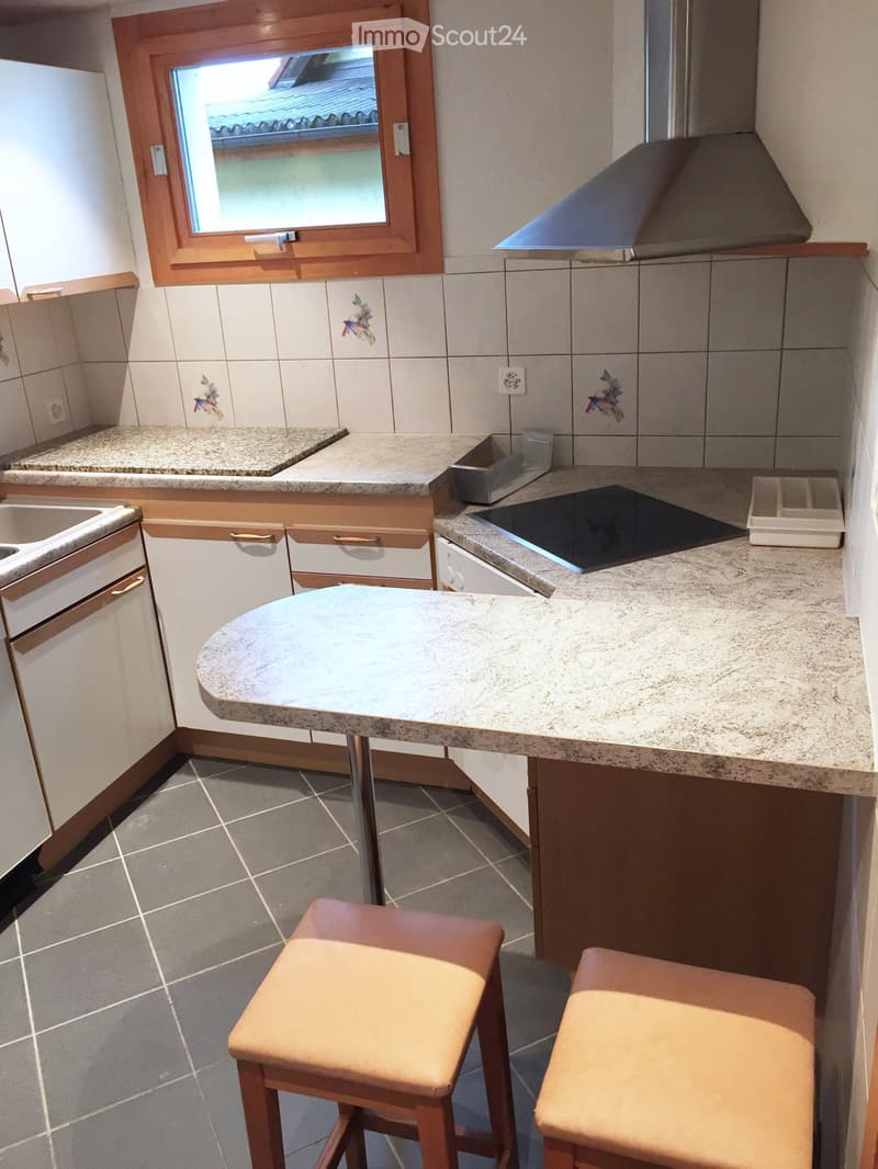 Modern kitchen with granite countertops, white cabinets, a round table with two stools, a sink, dishwasher, stove, and range hood. The room features tiled walls and a window with wooden framing.