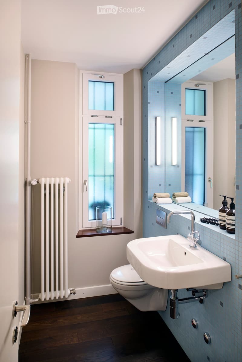 Modern bathroom with blue tiled walls, white sink, chrome faucet, toilet, and a large mirror. Features a radiator, a window with frosted glass, and a small shelf with a candle and glass.
