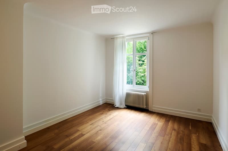 Empty room with wooden flooring, white walls, a window with white curtains, and a radiator below the window.