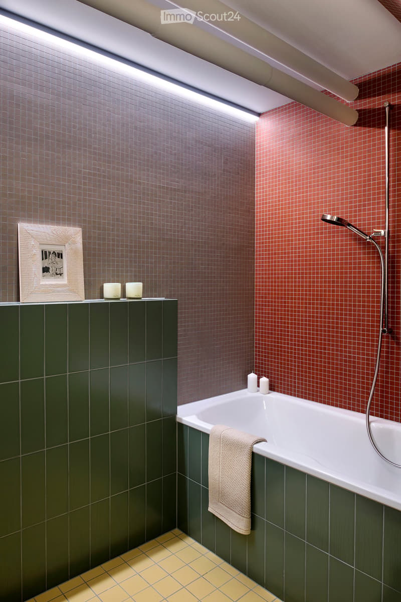 Modern bathroom with a white bathtub, green tiles, red mosaic tiles, and a towel draped over the edge. Includes a framed picture and candles on a shelf.