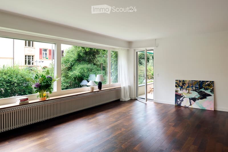 Bright, minimalist living room with wooden flooring, large windows, a radiator, a small table with a vase of flowers and books, and a colorful abstract painting on the wall.