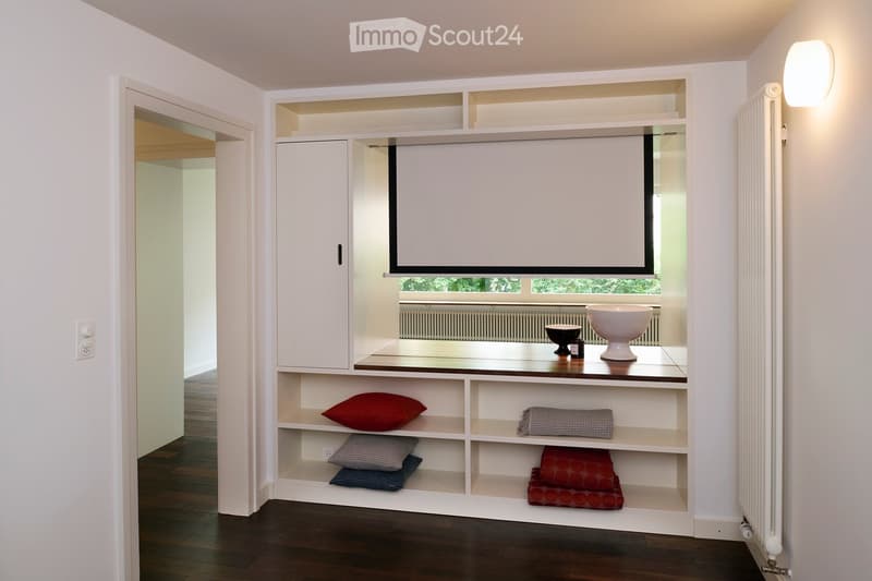 Modern, minimalist room with built-in white shelving and a projector screen. Features a dark wooden floor, white walls, and decorative pillows on the shelves. A small table holds a large white bowl and smaller decorative items.