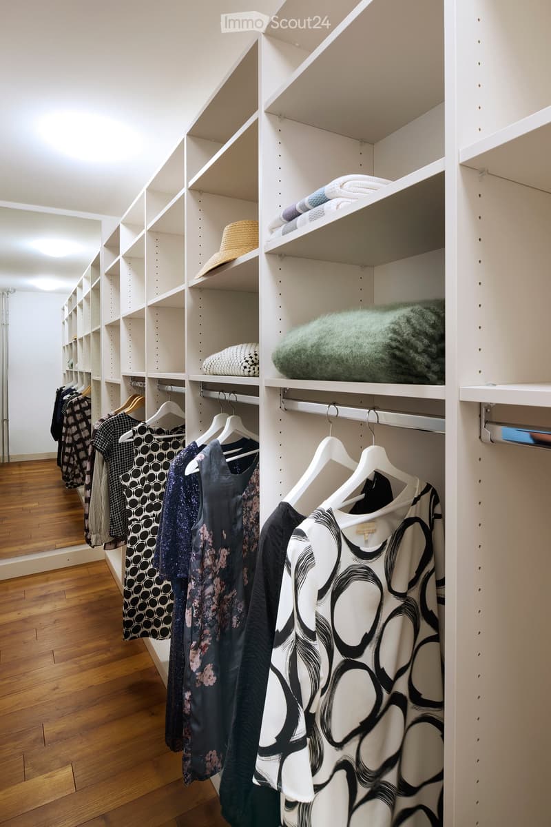 Well-organized walk-in closet with white built-in wardrobe and shelves, containing neatly arranged clothing, a hat, and a green blanket.