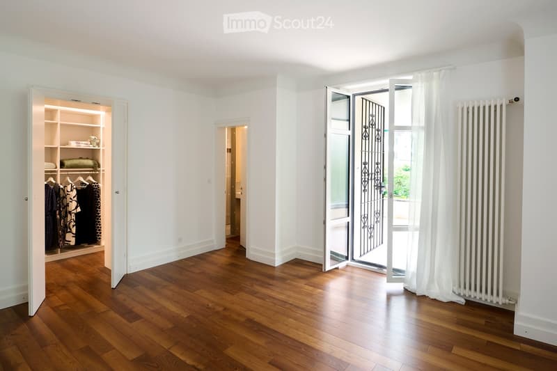 Empty room with wooden flooring, white walls, built-in closet with shelves, and a large window with white curtains.