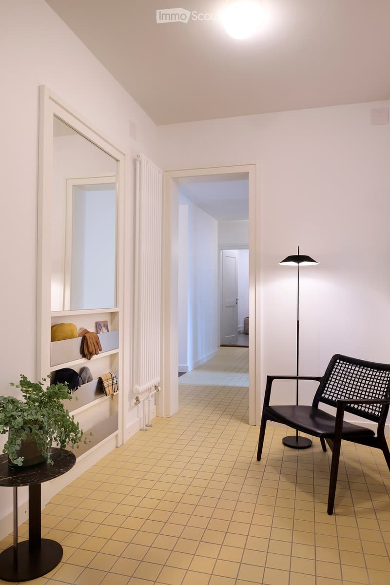 A modern hallway with a black woven chair, a round black table with a potted plant, a shelf with folded blankets and gloves, and a tall floor lamp.