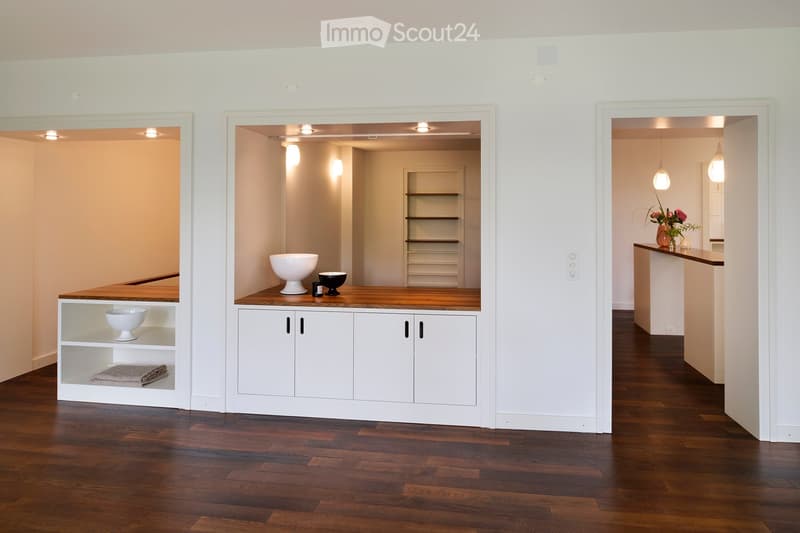 A modern hallway with three distinct areas: a small seating area with a wooden bench and white bowls, a central area with a wooden counter and white cabinets, and a right-side area with a wooden counter and a vase with flowers. The floor is made of dark h