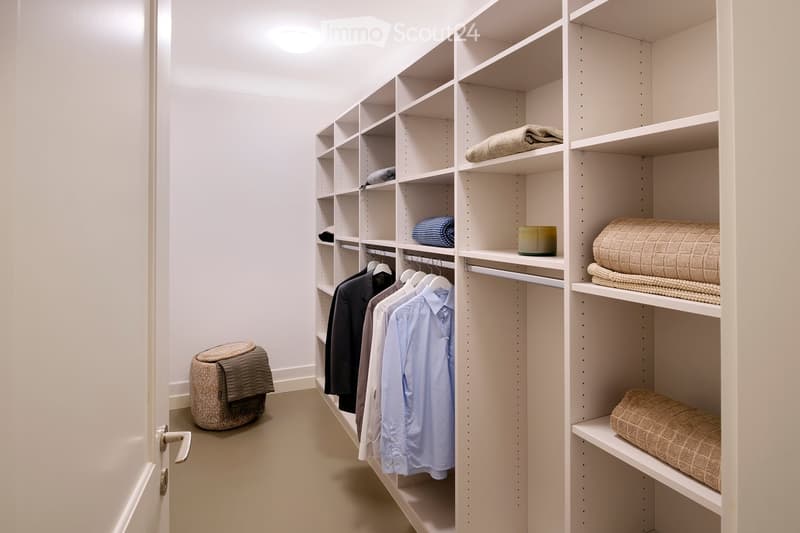 A well-organized walk-in closet with white built-in wardrobes and shelves, neatly arranged clothing, folded blankets, and a small stool.
