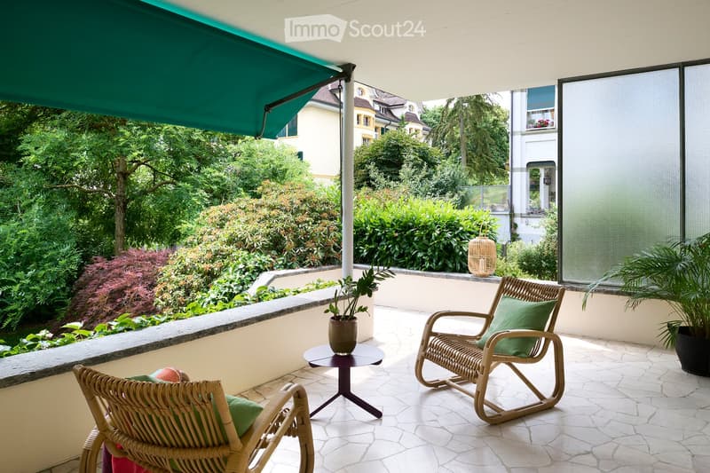 A well-maintained balcony with two wicker chairs, a small round table, and potted plants, featuring a green umbrella and a view of a lush garden.