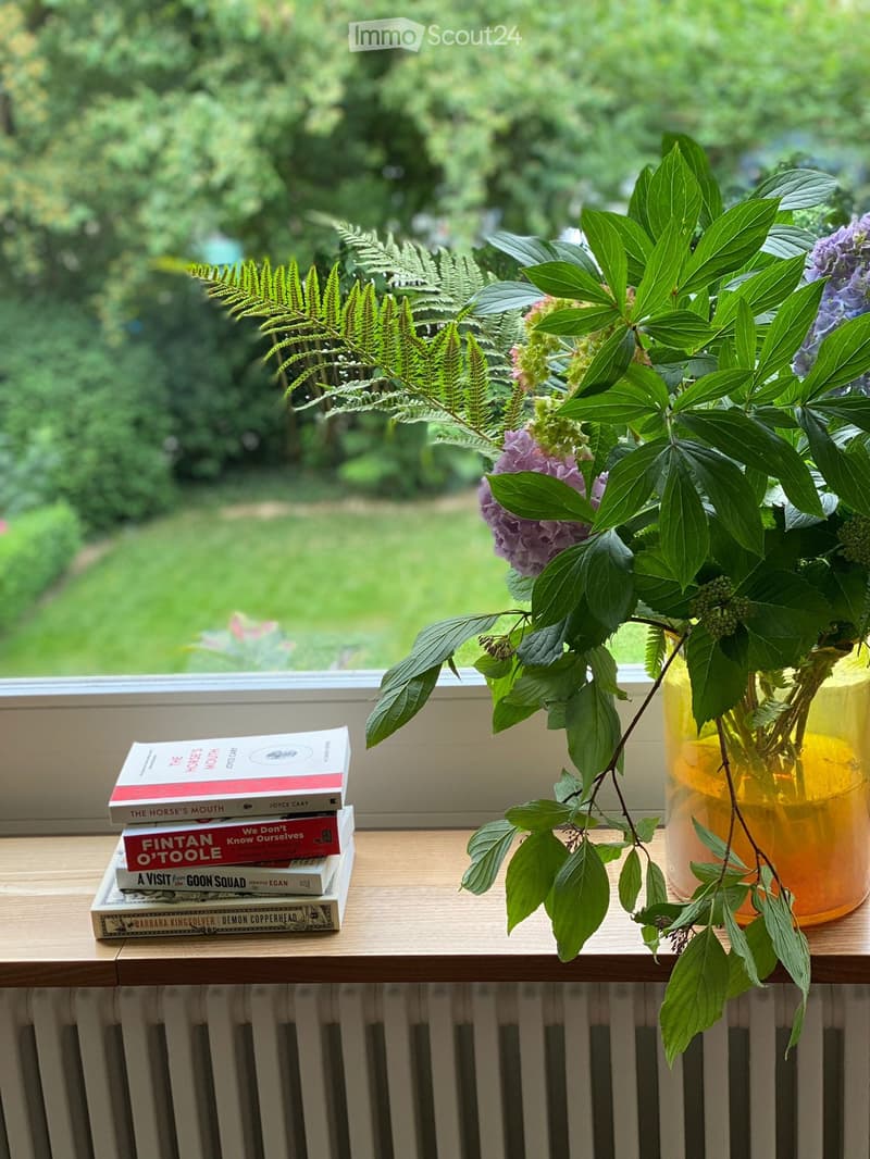 A window sill with a vase of flowers and a stack of books, overlooking a green garden area.