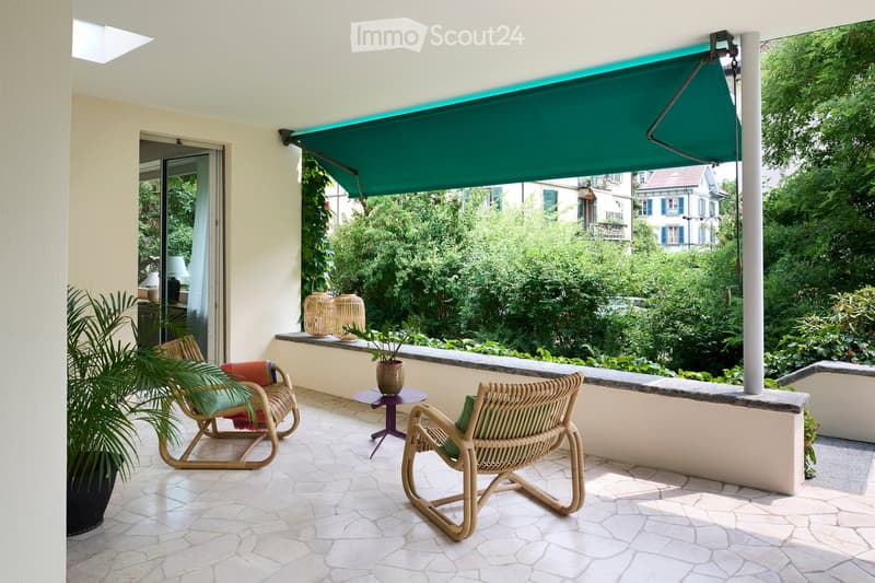Balcony with a green awning, two wicker chairs, a small table, potted plants, and wicker baskets on the ledge.