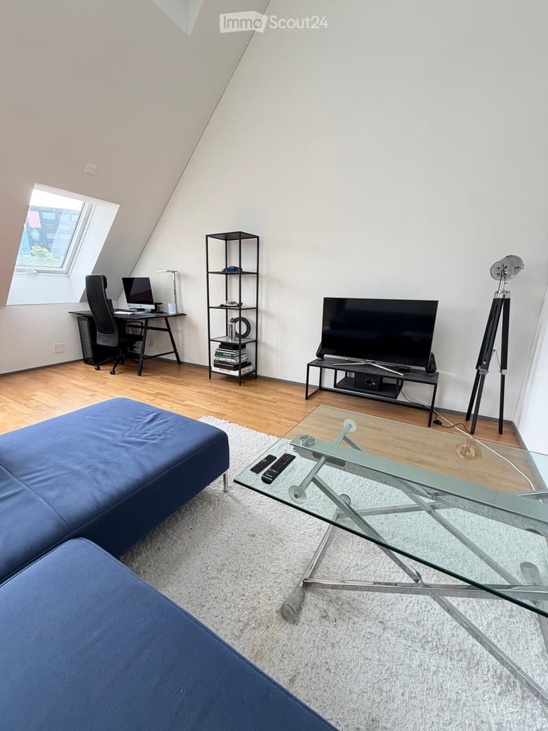Modern living room with a blue sofa, glass coffee table, TV, desk with chair, and shelving unit.