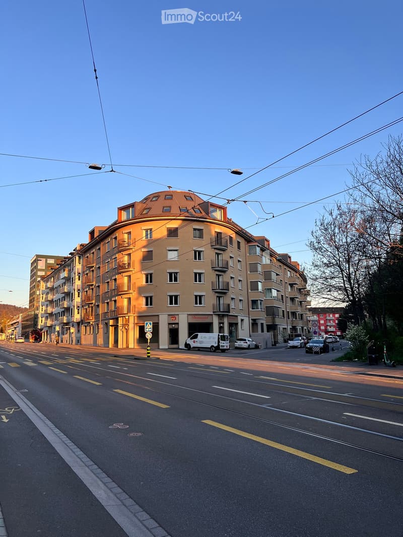 Multi-story building with a rounded facade, balconies, and large windows, located on a street with parked cars and traffic markings.