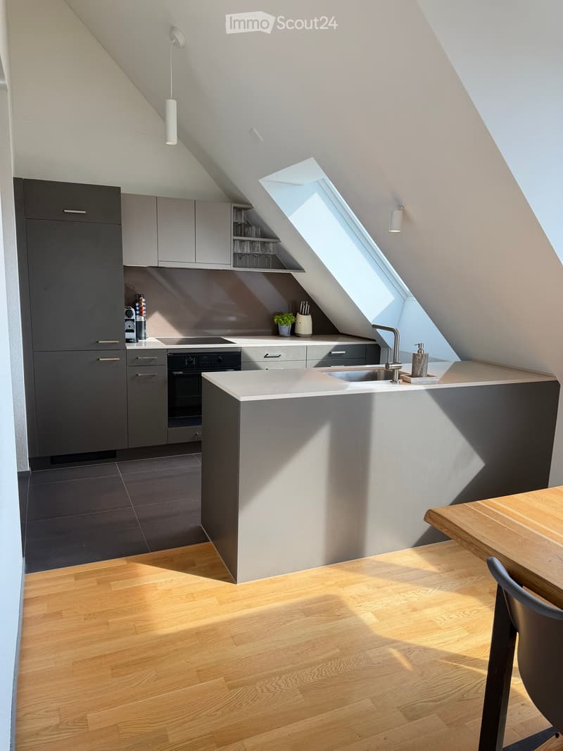 Modern kitchen with grey cabinets, white countertops, a central island, and a skylight window. Includes a sink, faucet, oven, and a small dining area with a wooden table and chair.