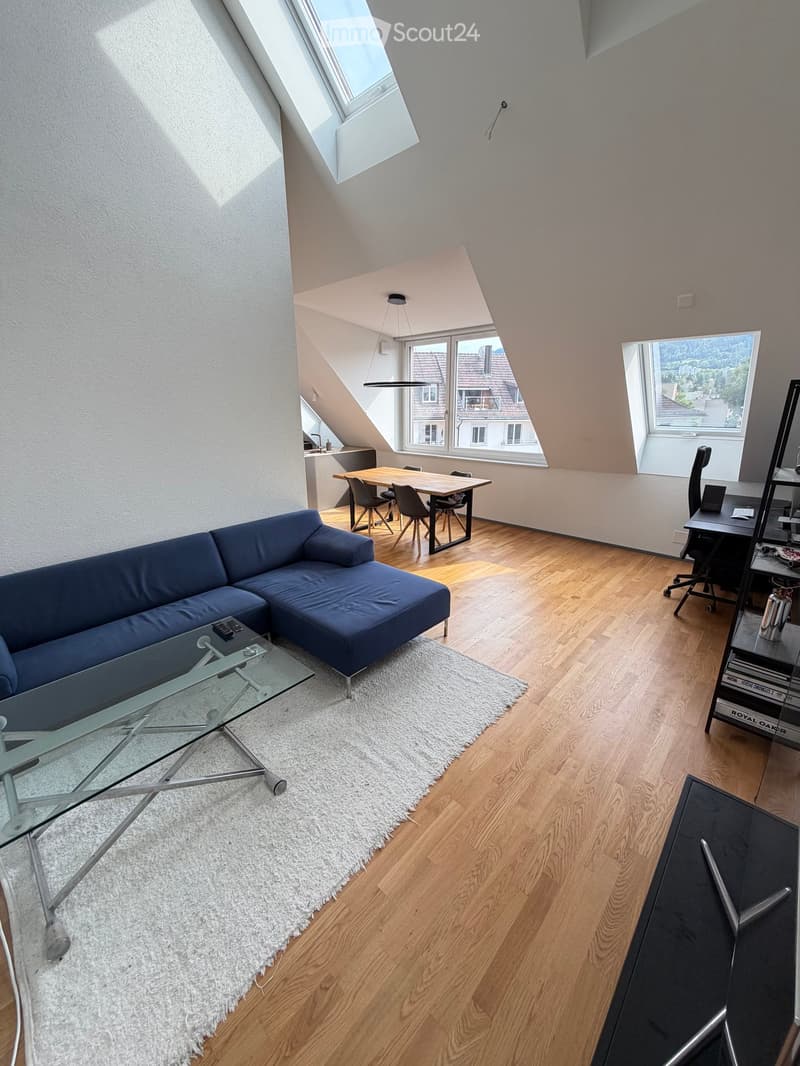 Bright, open living space with wooden flooring, large windows, and skylights. Features a blue sectional sofa, glass coffee table, dining table with chairs, desk with laptop, and bookshelf.