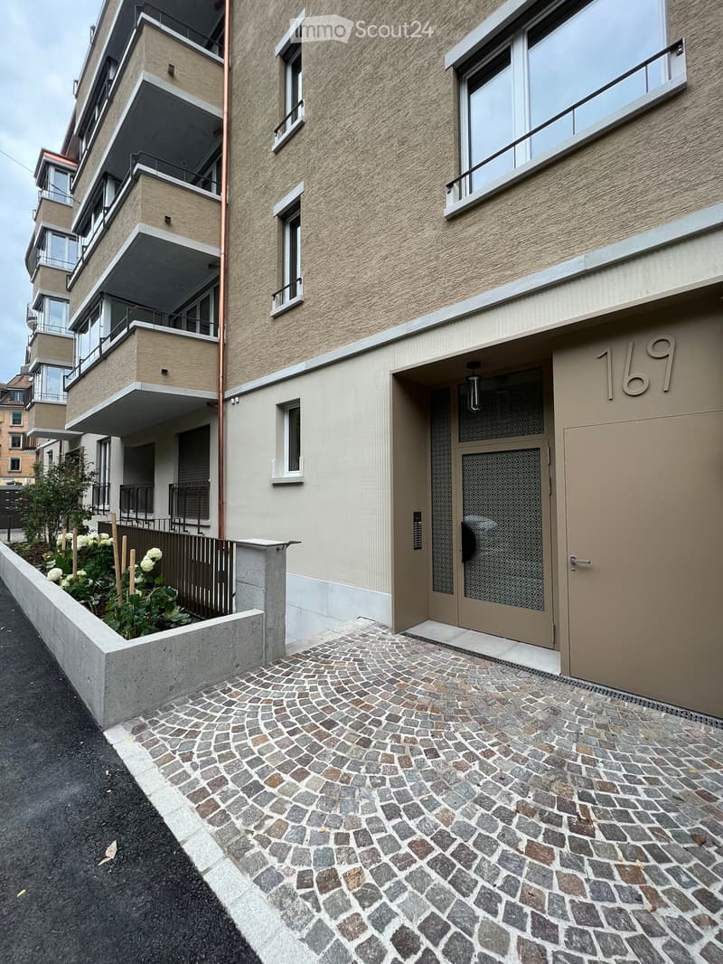 Modern multi-story building with beige facade, large windows, balconies, and a cobblestone pathway leading to the entrance.