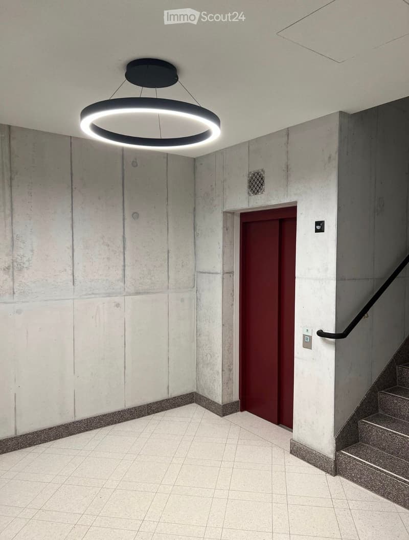 Modern hallway with white tiled floor, grey concrete walls, a red door, a staircase with a black handrail, and a circular pendant light fixture.