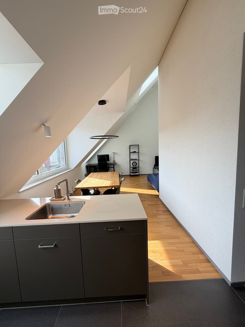 Modern kitchen with grey cabinets, a sink, and a dining area with a wooden table and chairs. There is a computer desk and a blue sofa in the adjacent space.