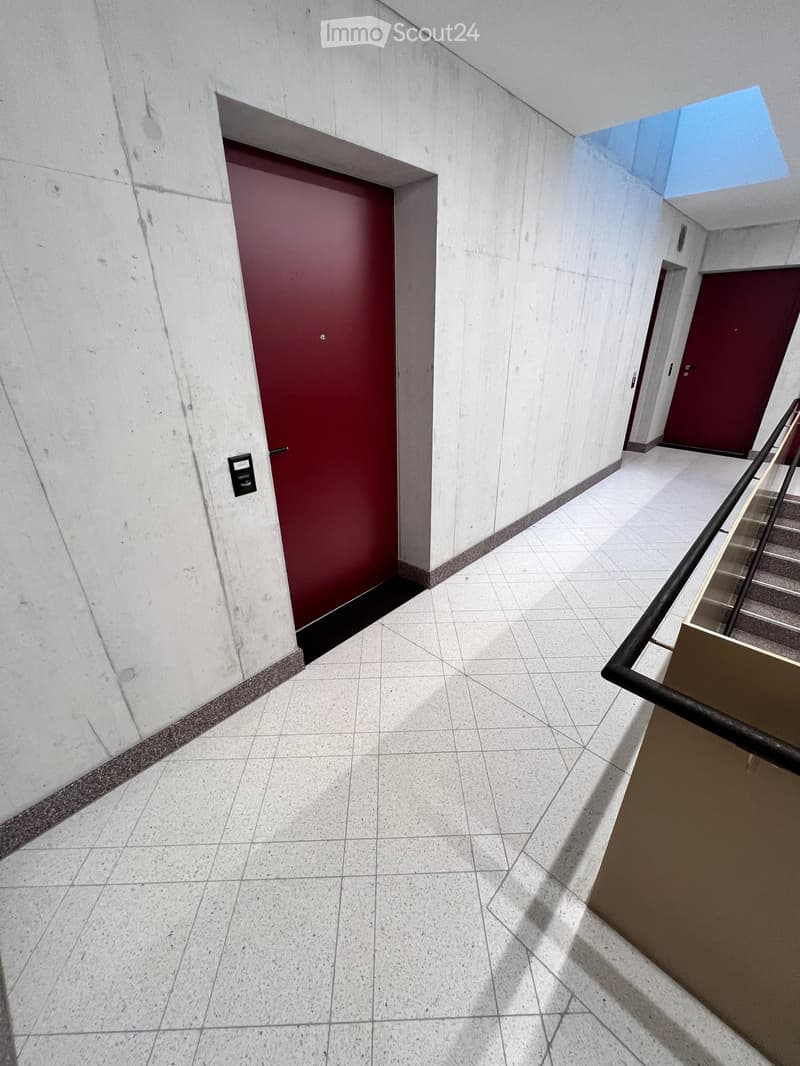 Modern hallway with white tiled floor, light grey walls, and red doors. Features a staircase with a black railing on the right side.