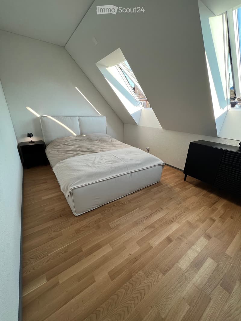 Modern bedroom with wooden flooring, a large bed with white bedding, nightstand with lamp, and a black cabinet.