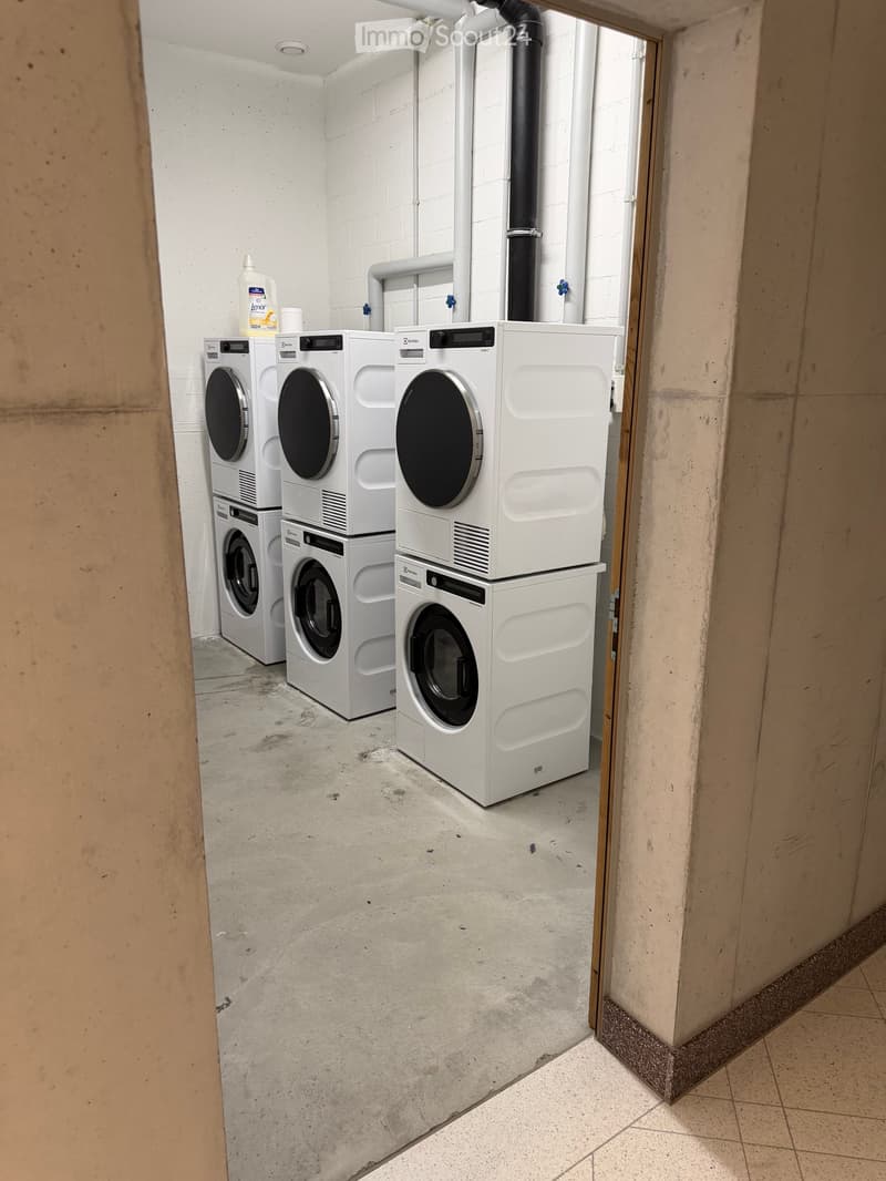 A small, clean laundry room with three stacked washing machines and dryers, white walls, and a concrete floor.
