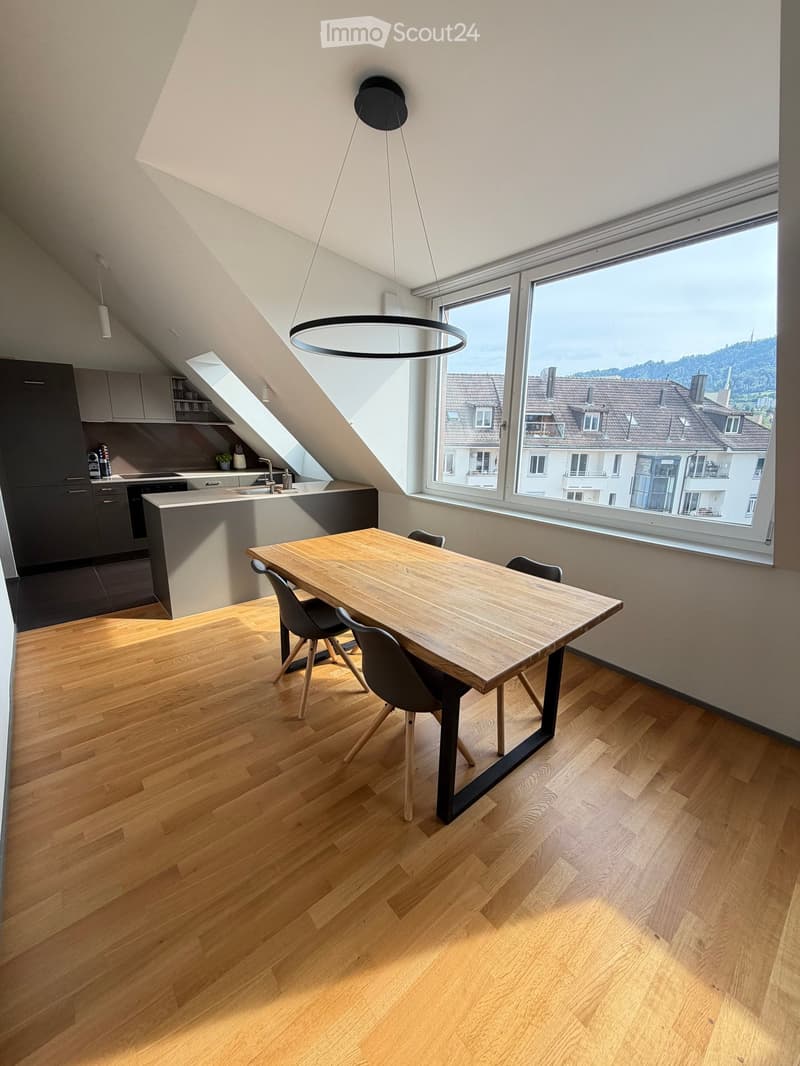 Modern, open-plan dining and kitchen area with wooden flooring, large windows offering a view, a wooden dining table with four chairs, and a sleek kitchen area with cabinets and a sink.