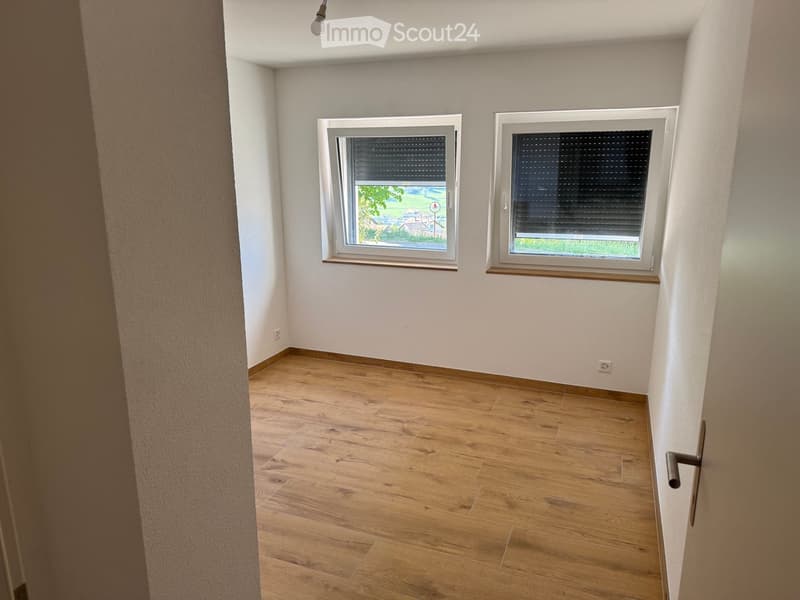 Empty room with wooden flooring, white walls, and two windows with blinds. Natural light streams in from the windows, and the room has a simple, clean design with no furniture or decorations.
