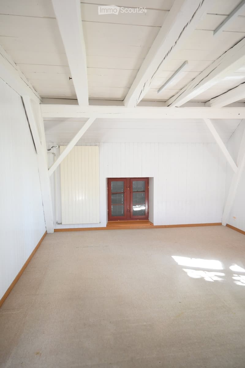 Empty room with white wooden walls, exposed wooden beams, and a single window with red frame. The floor is carpeted in light beige.