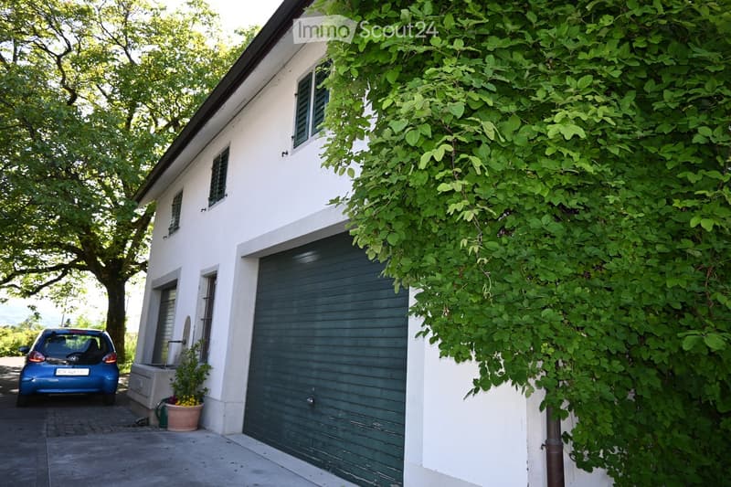 White house with a garage, covered in green ivy, with a small garden area and a blue car parked nearby.