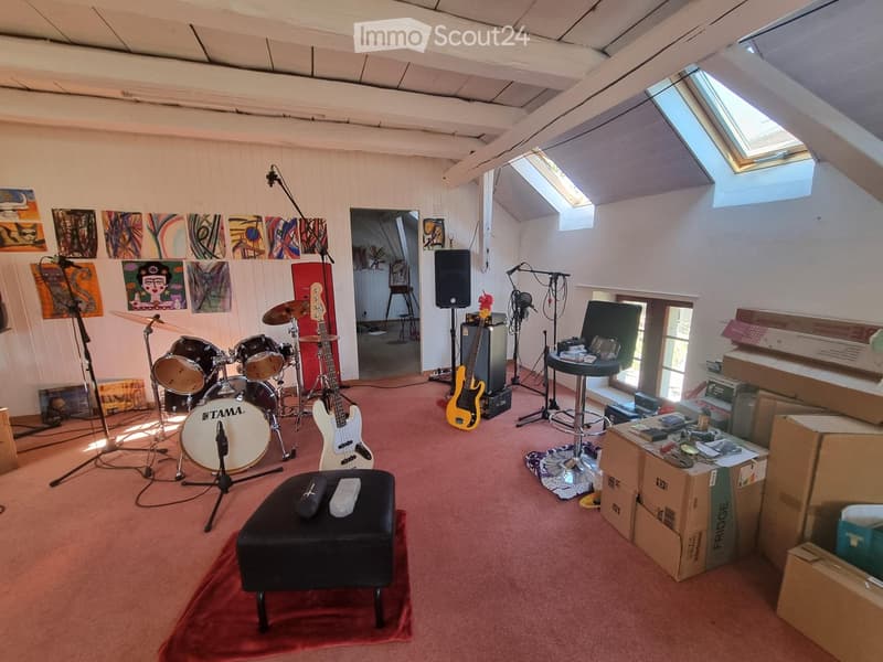 A spacious room with a drum set, guitars, speaker, microphone stands, and artworks on the walls. The room has wooden beams on the ceiling and skylights providing natural light.