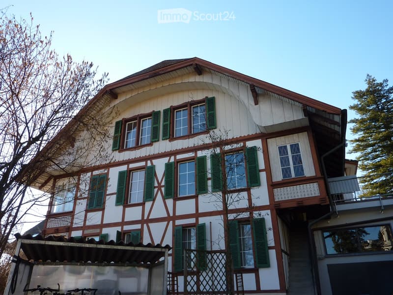 Two-story house with white and brown exterior, green shutters, multiple windows, and a balcony on the upper level.
