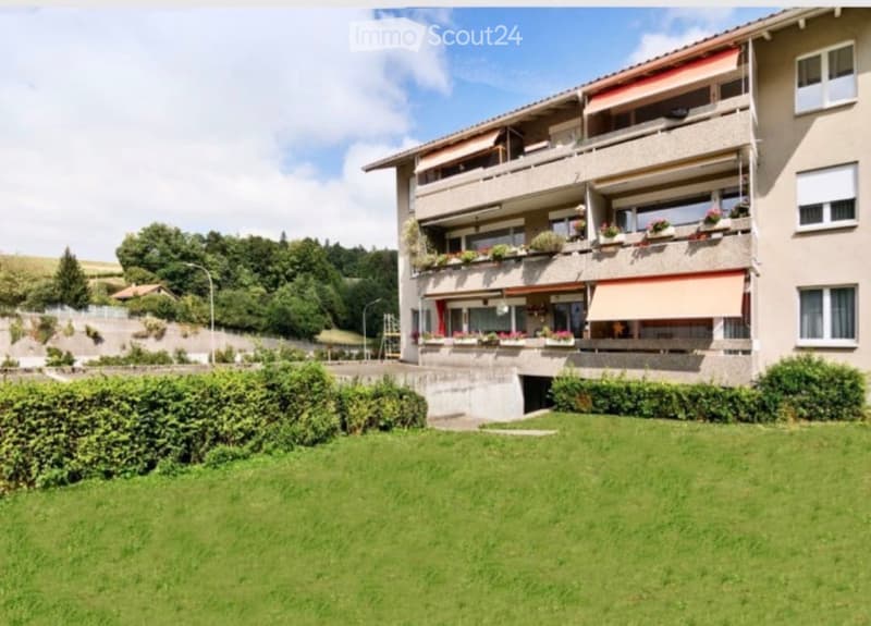 Multi-story apartment building with balconies, greenery, and a well-maintained lawn in front.