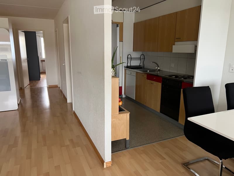 A modern kitchen with light wooden cabinets, stainless steel appliances, a white dining table, and a black chair. The floor is made of light wooden planks.