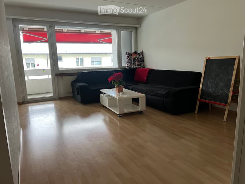 Modern living room with wooden flooring, large window, black sofa, white coffee table, potted plant, and blackboard.