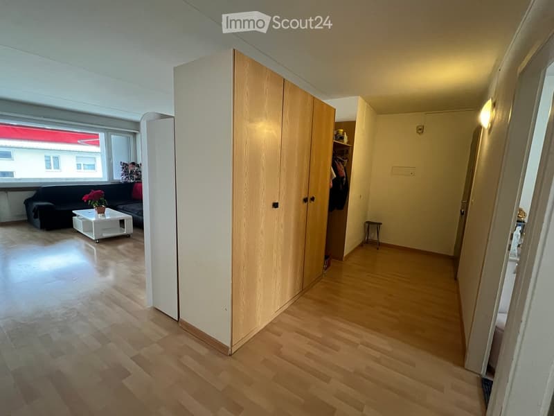 A furnished room with a sofa, coffee table, flower pot, wardrobe, and a small stool. The room has wooden flooring and large windows letting in natural light.