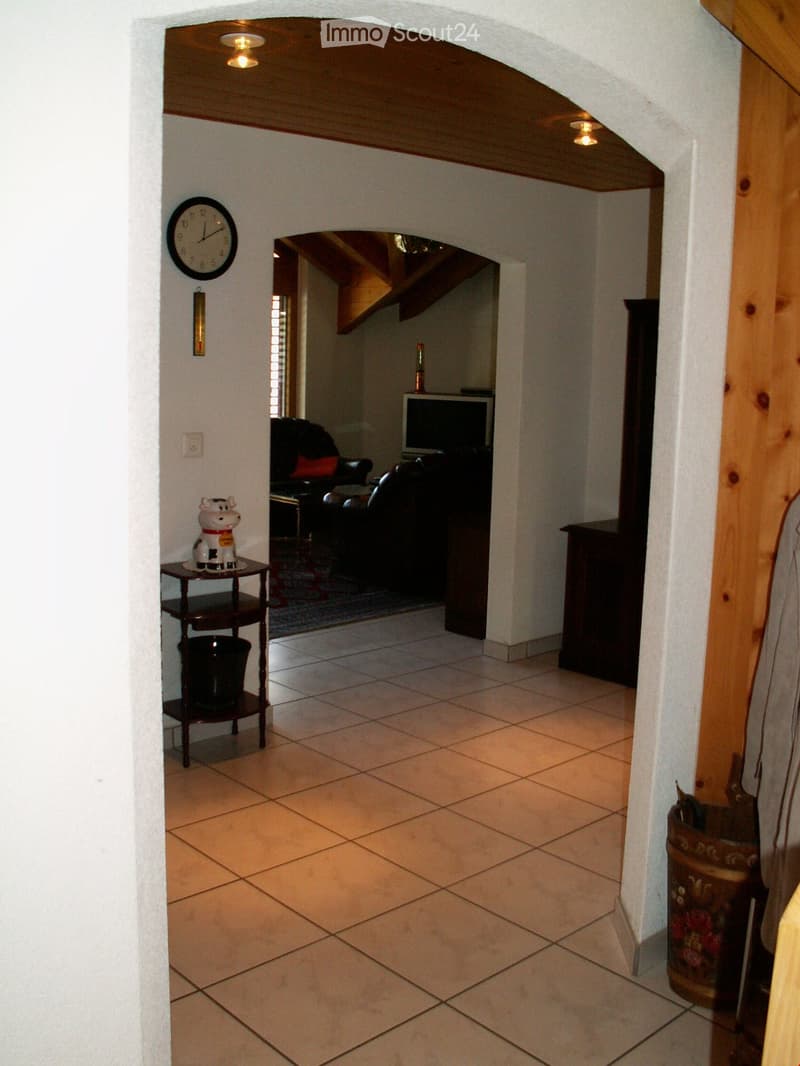 Tiled floor, ceiling lamps, arched entrance, small table with decorative cow, wall clock, brown wooden cabinet, living room with sofa and TV