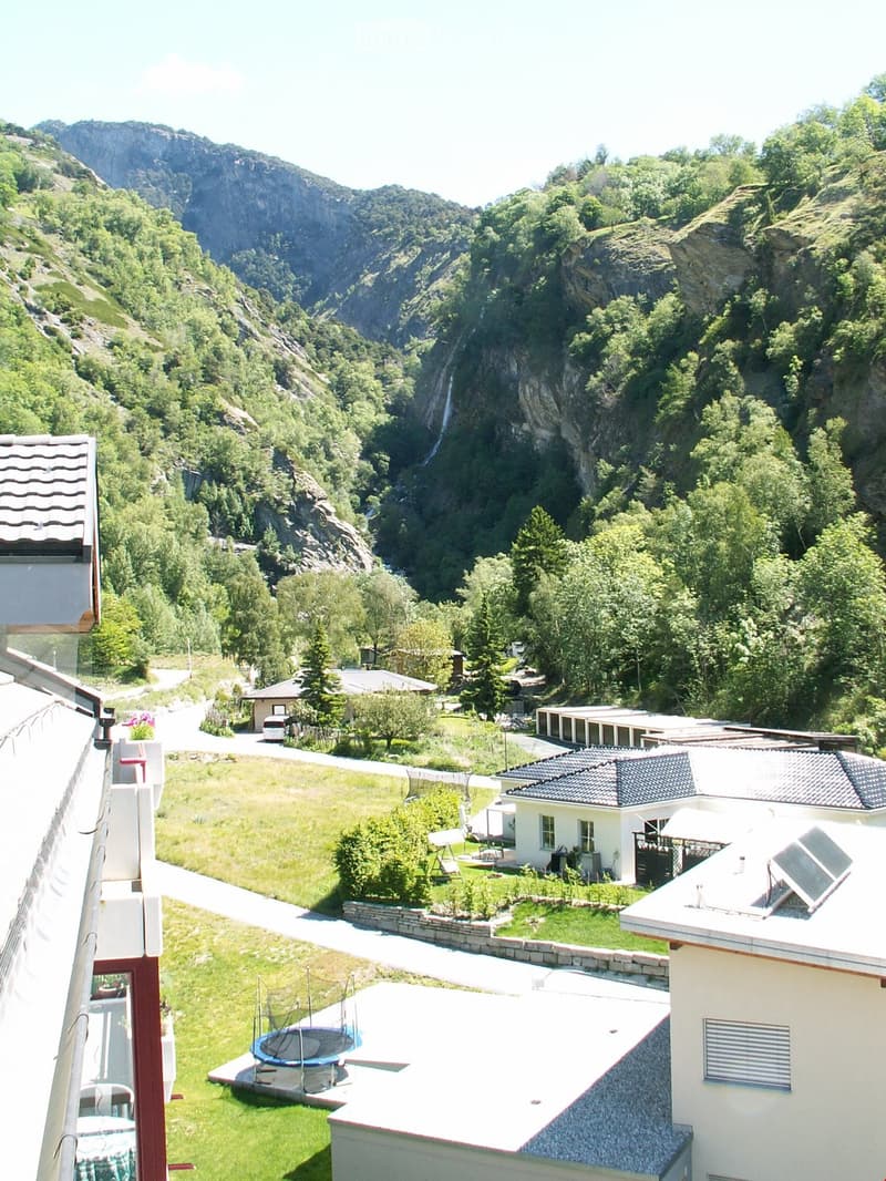 The aerial view of a building showcases a picturesque landscape, including a mountain, waterfall, grass, and a car parked on the road. The building features a roof with solar panels, a table, and a trampoline.