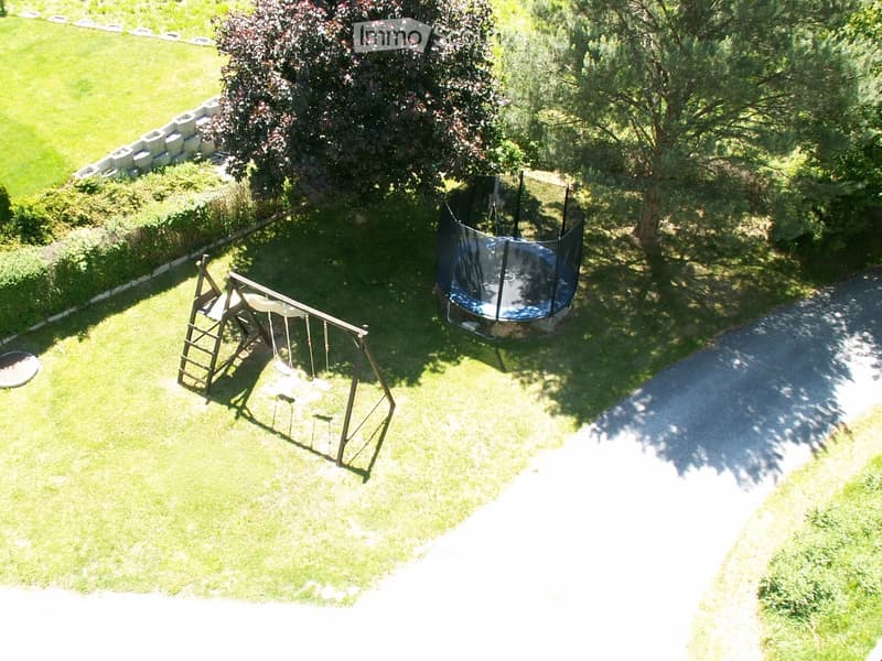 a large grass field, there is a swing, a trampoline, and a retaining wall