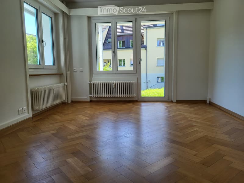 Empty room with wooden herringbone flooring, large windows, radiators, and a sliding glass door leading to a balcony.