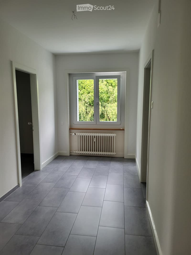 Empty hallway with tiled floor, white walls, a window with a radiator below, and two doors on either side.