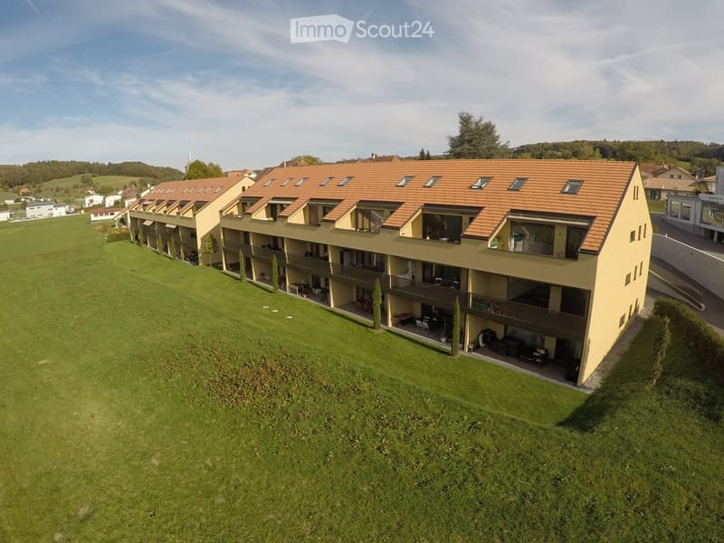 Apartment building with multiple floors, balconies, and a brown roof, set in a large green field