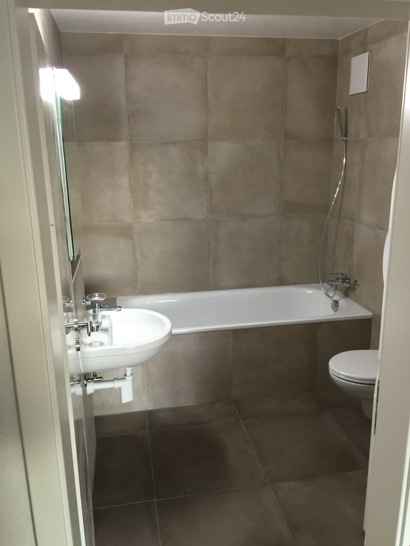 Modern bathroom with a white sink, bathtub, toilet, and a glass shower enclosure. The walls are tiled in a neutral tone, and the floor is covered with dark tiles.
