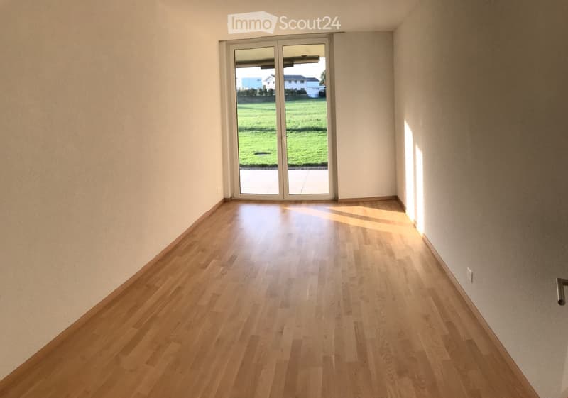 Empty room with wooden flooring and large sliding glass door leading to a green outdoor area.