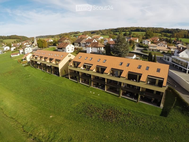 Aerial view of a modern multi-story residential building with orange-tiled roofs and balconies, surrounded by green lawns and neighboring houses in a rural setting.