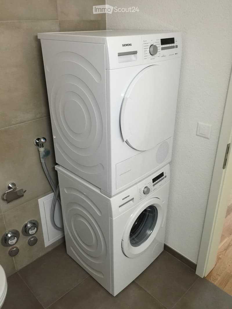 A compact bathroom with a stacked Siemens washing machine and dryer unit, tiled walls, and a wooden floor visible in the background.