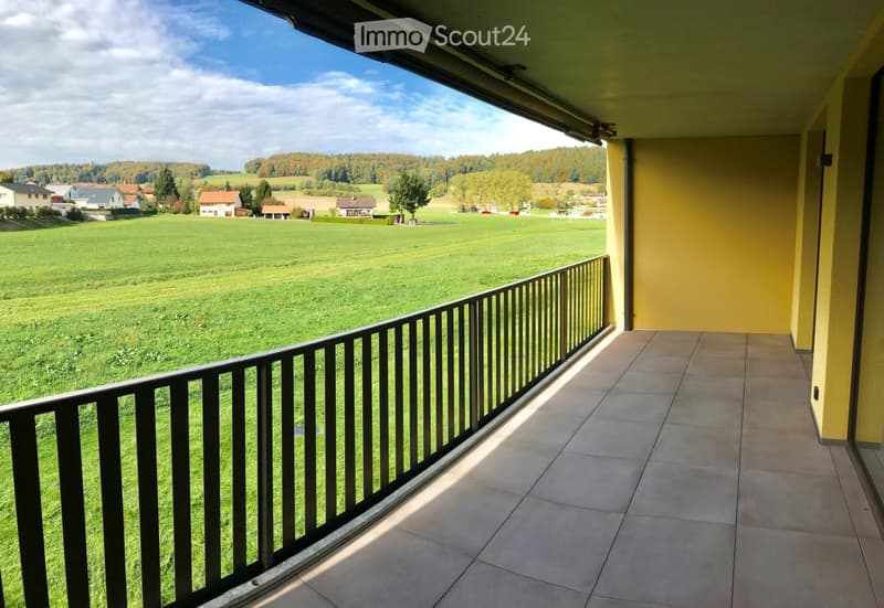 Balcony with tiled floor, black metal railing, overlooking a green field and rural houses.